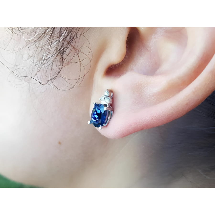 Oval blue sapphire stud earring with three small diamonds in 14K white gold on an ear.