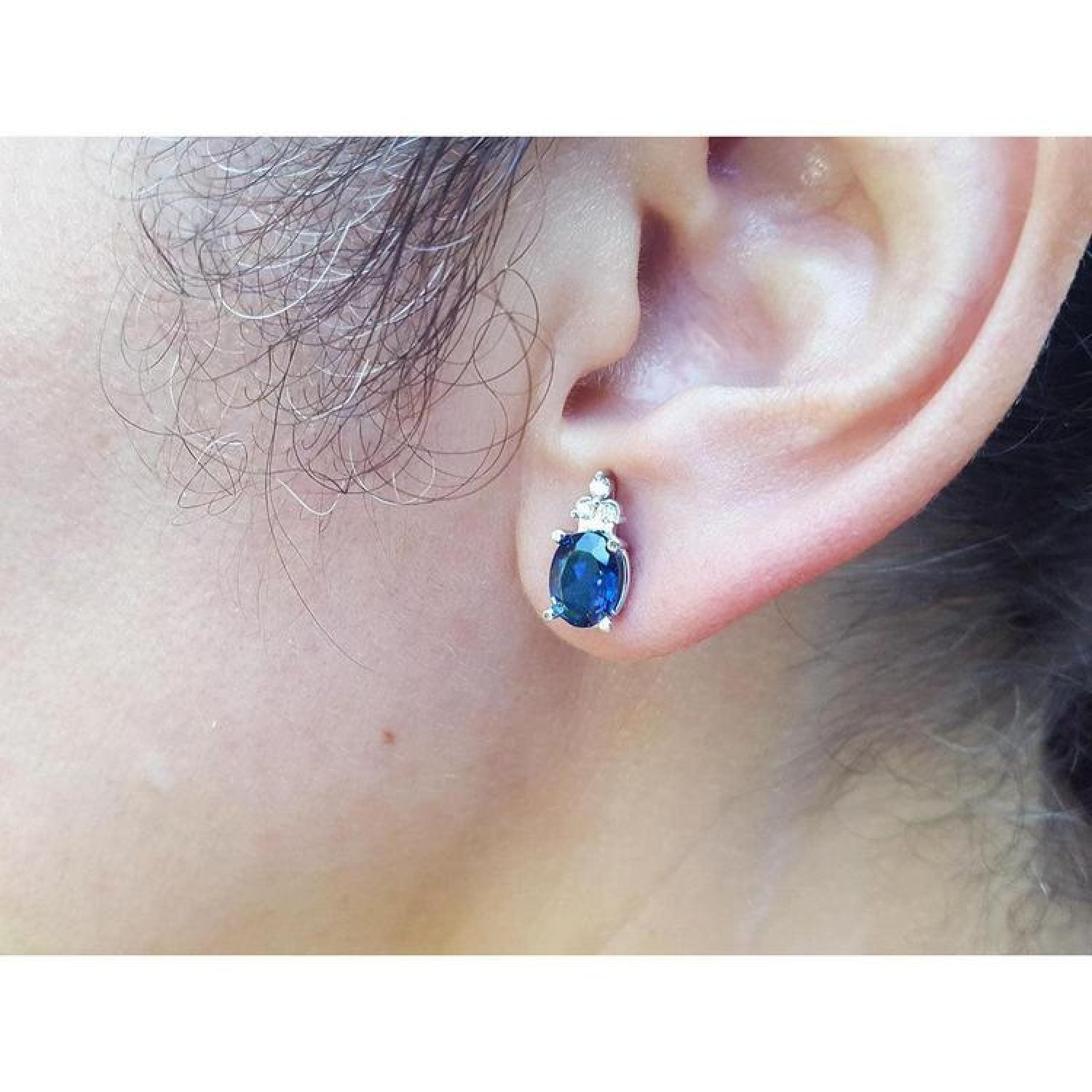 Oval blue sapphire stud earring with three small diamonds, set in 14K white gold on an ear.
