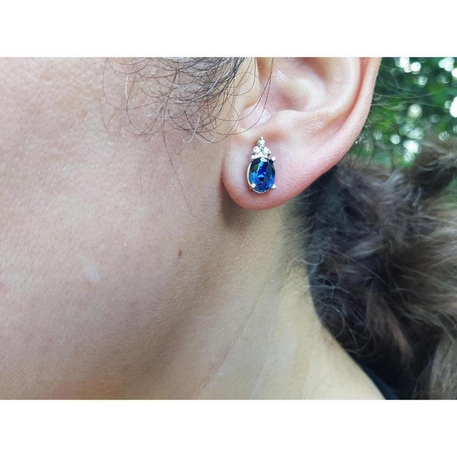 Oval blue sapphire stud earrings with three prong-set diamonds in 14K white gold on an ear.