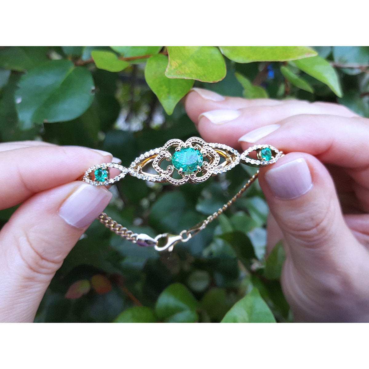 Gold bracelet with real colombian emeralds held between fingers against a leafy background.