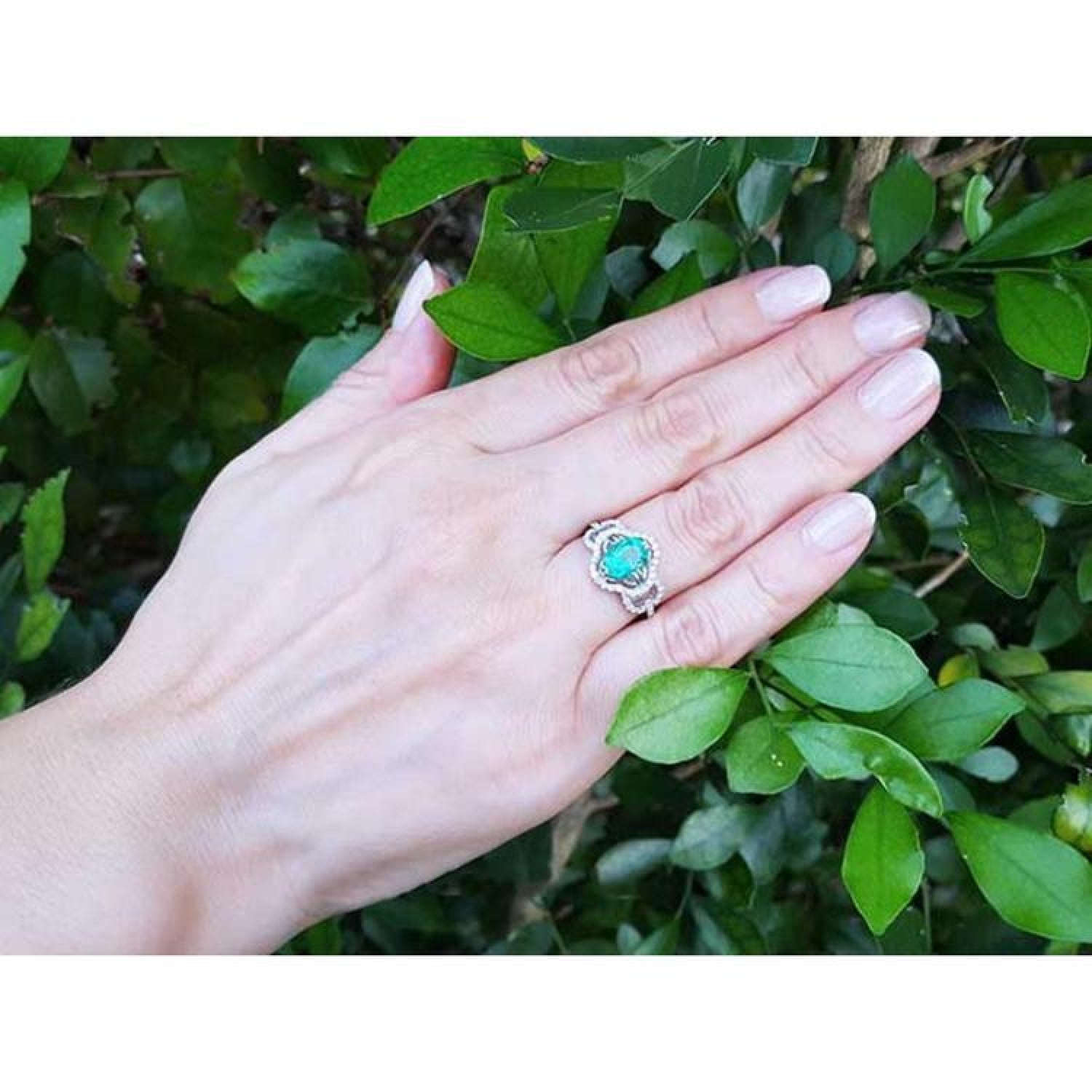 Oval emerald clover ring with diamond halo in 14K white gold on a hand with green leaves.