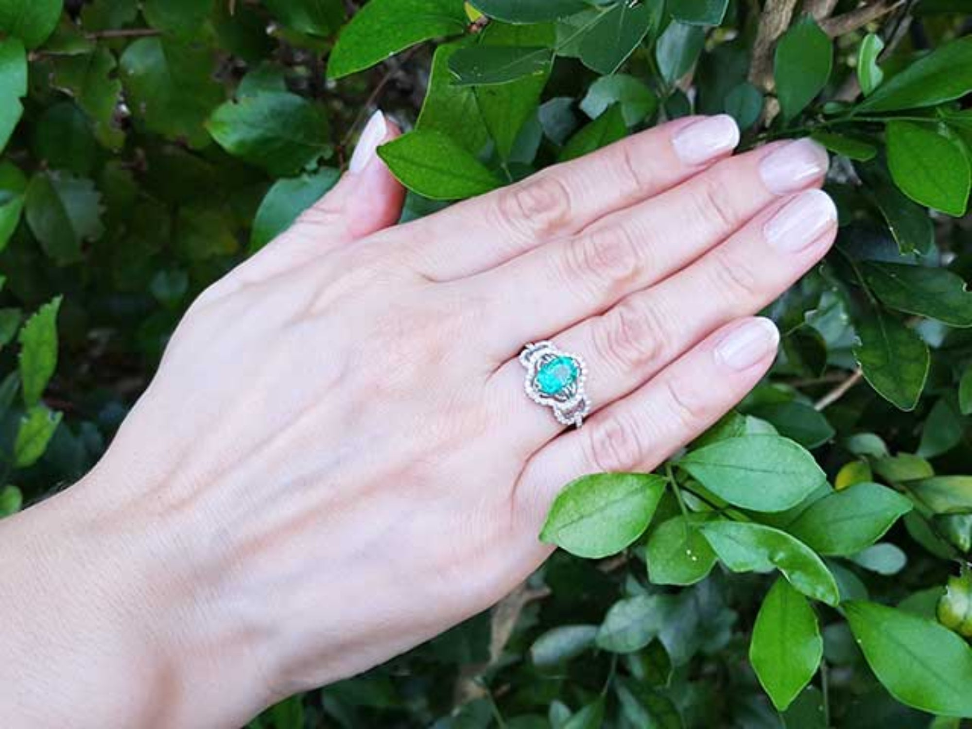 Hand wearing an oval cut emerald ring with accent diamonds against a green leafy background