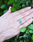 Hand wearing an oval cut emerald ring with accent diamonds against a green leafy background