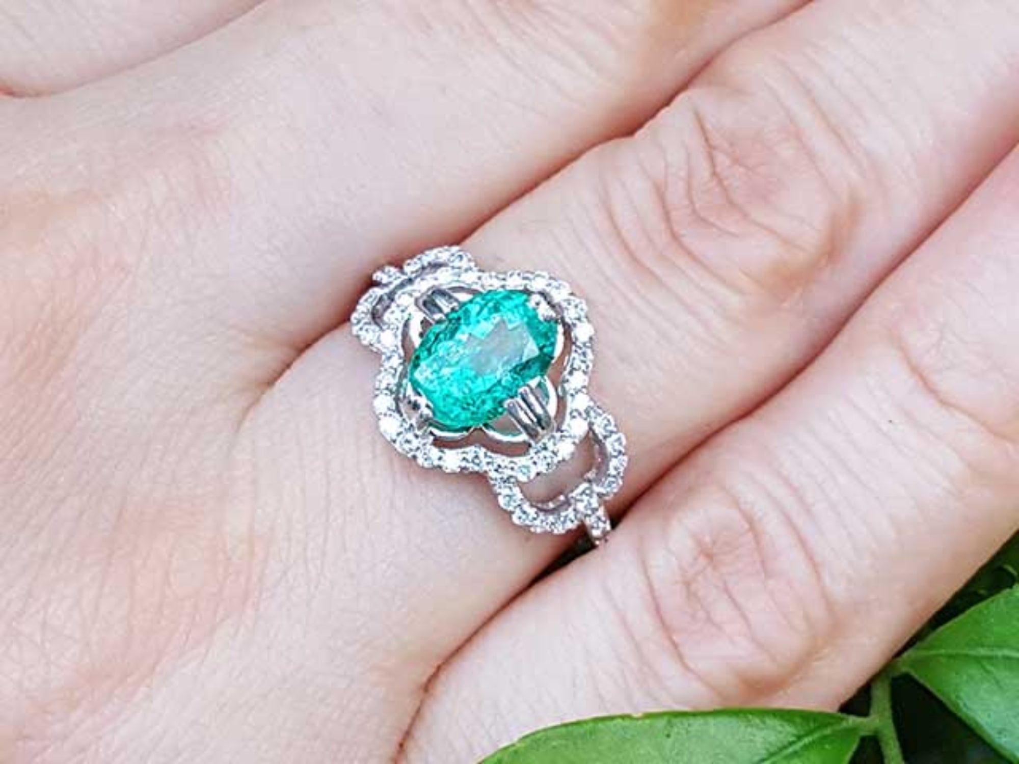 A 14k white gold clover ring with an oval cut emerald surrounded with natural diamonds on a person&#39;s finger.