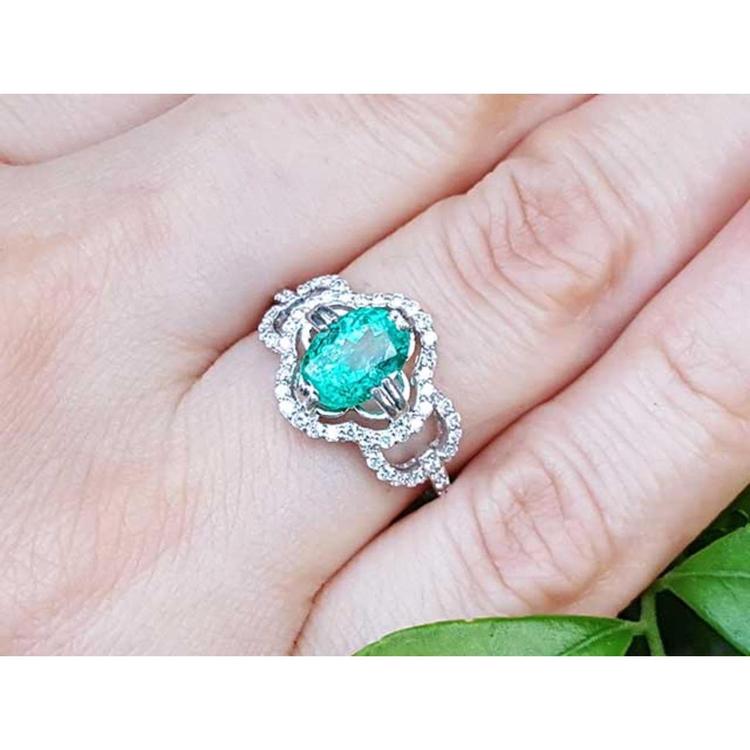 A 14k white gold clover ring with an oval cut emerald surrounded with natural diamonds on a person's finger.