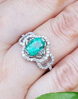 A 14k white gold clover ring with an oval cut emerald surrounded with natural diamonds on a person's finger.