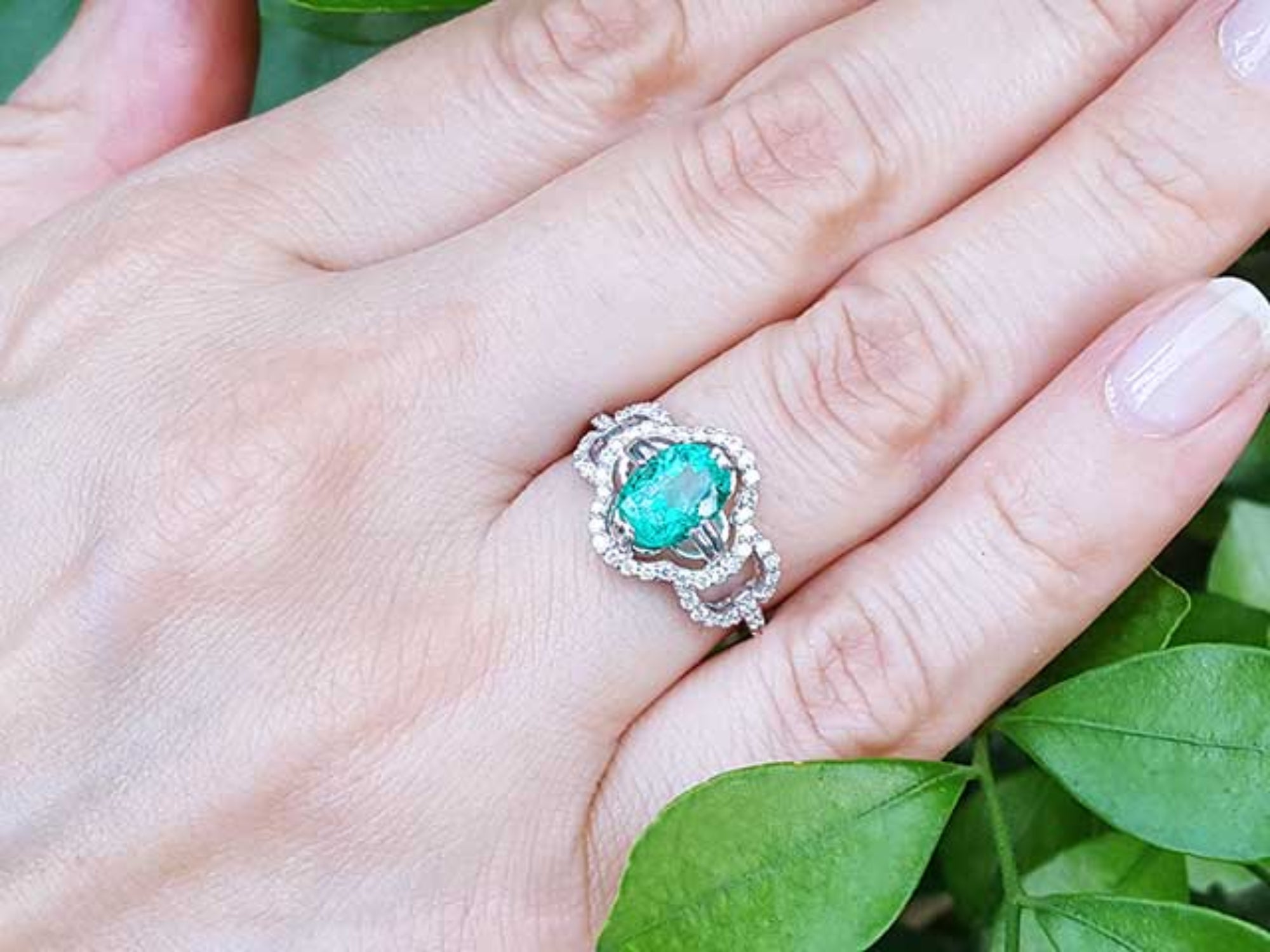 A clover ring with an oval cut emerald on a hand against a green leafy background.