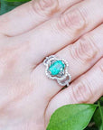 A clover ring with an oval cut emerald on a hand against a green leafy background.