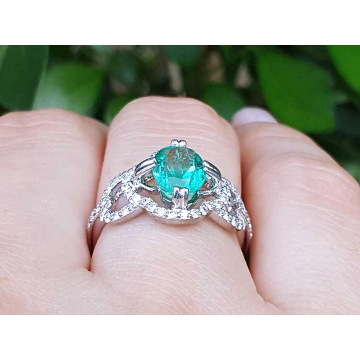 A white gold emerald ring on a finger against a blurred green leafy background.
