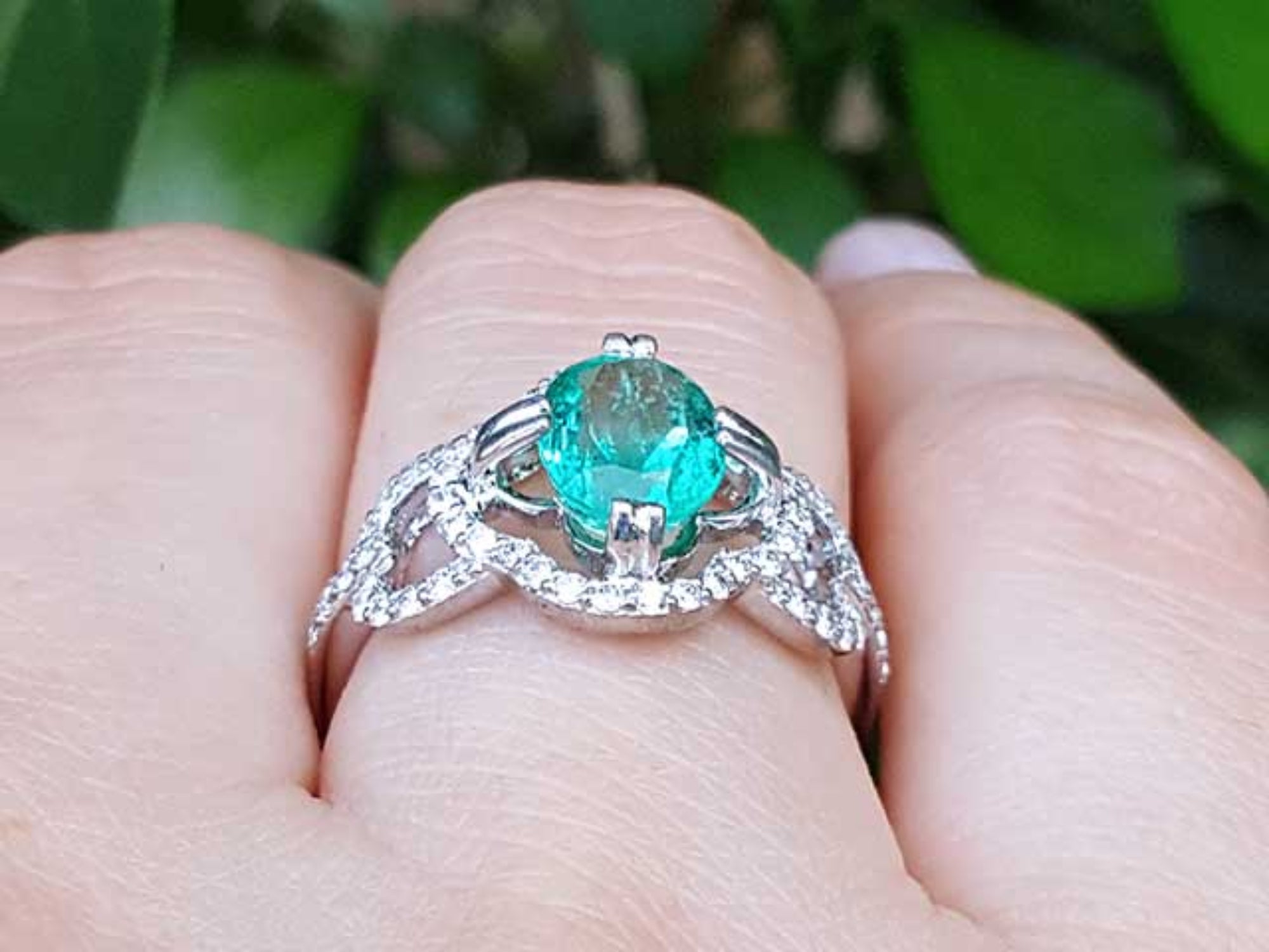 A white gold emerald ring on a finger against a blurred green leafy background.