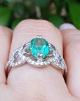 A white gold emerald ring on a finger against a blurred green leafy background.