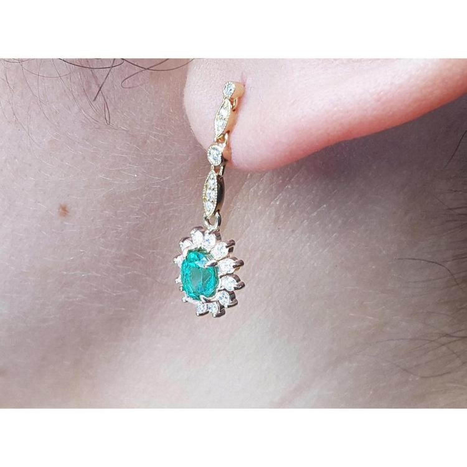 Oval emerald and halo diamond dangle earrings in 14K yellow gold, worn on an ear.