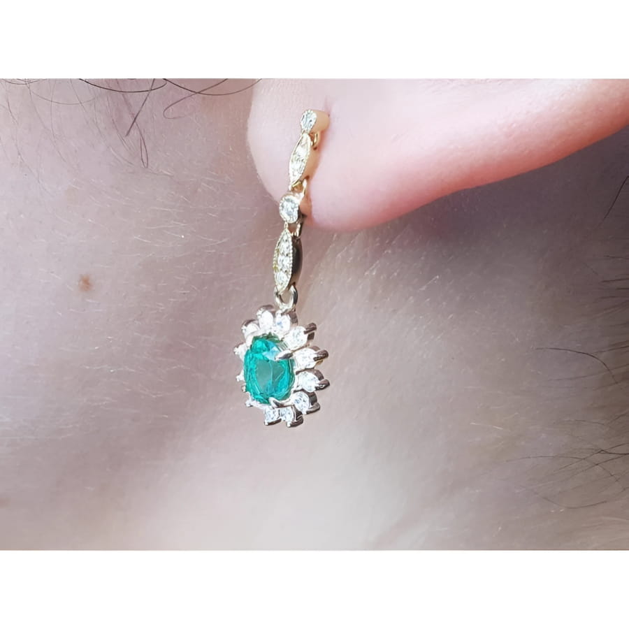 Oval emerald and halo diamond dangle earrings in 14K yellow gold, worn on an ear.