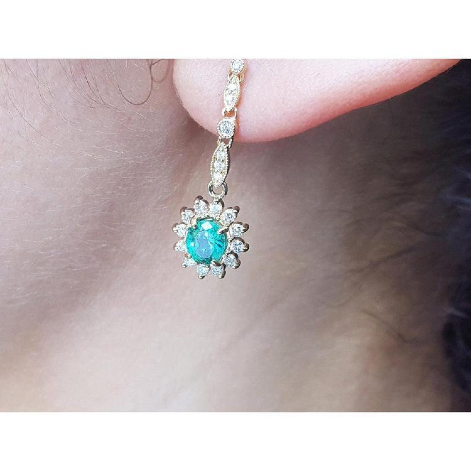 14K yellow gold dangle earring with oval emerald surrounded by halo diamonds on an ear.