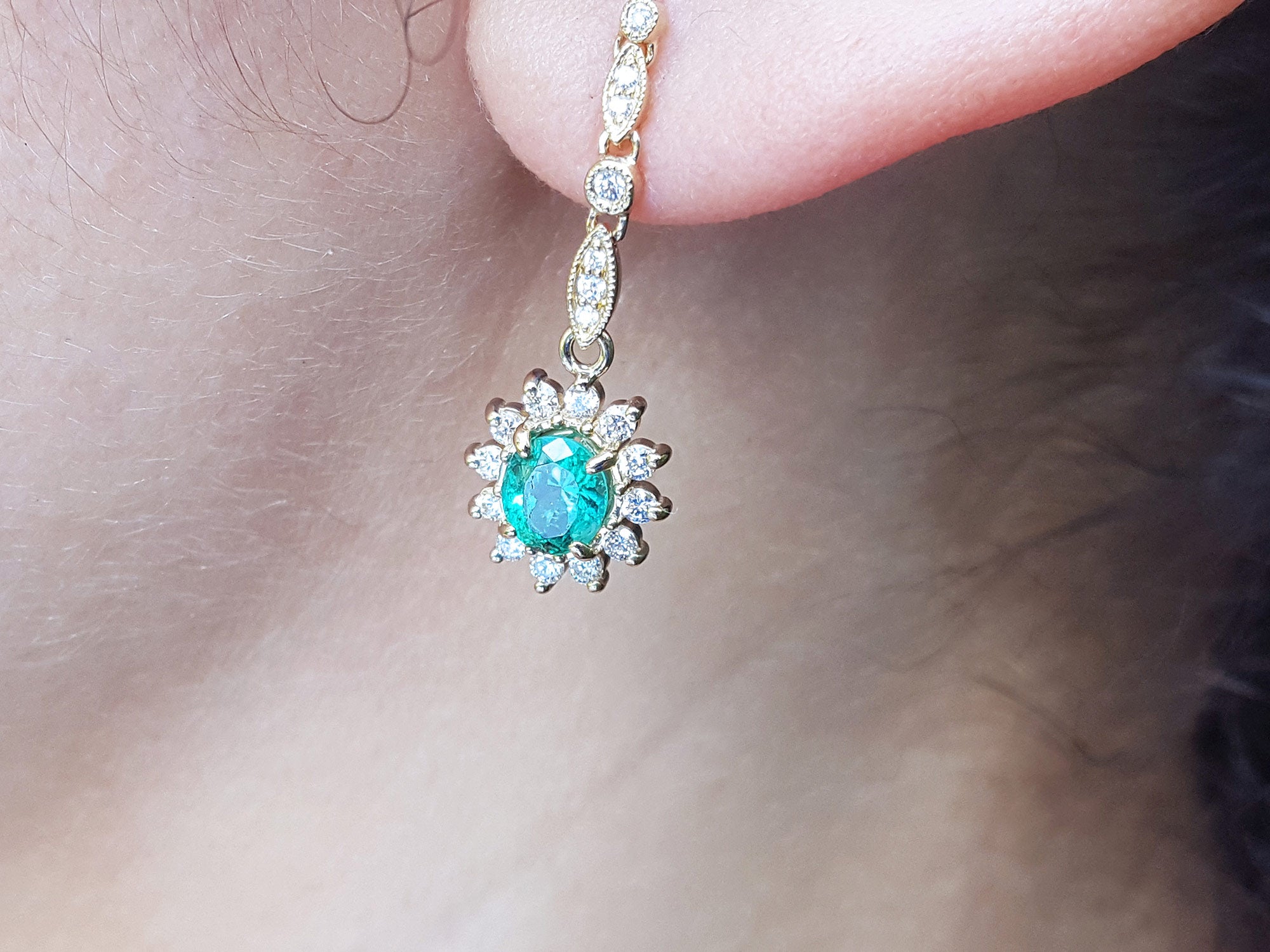 Close-up of an oval cut emerald encircled by a halo diamond earring on an ear. 