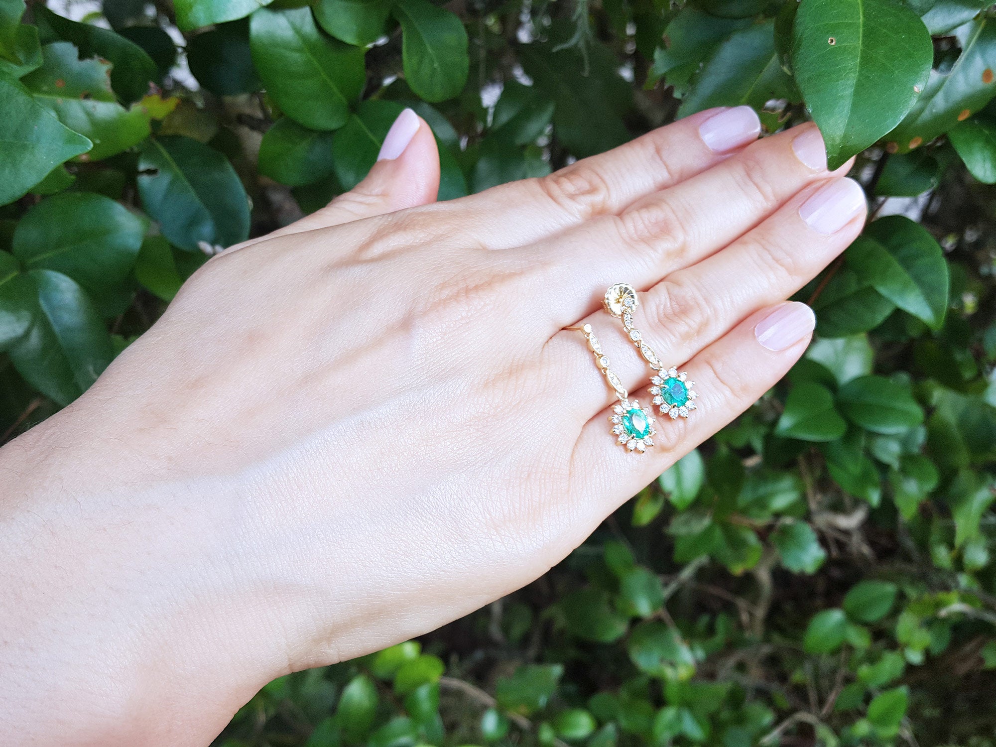 Hand displaying a pair of emerald earrings oval cut against a green leafy background