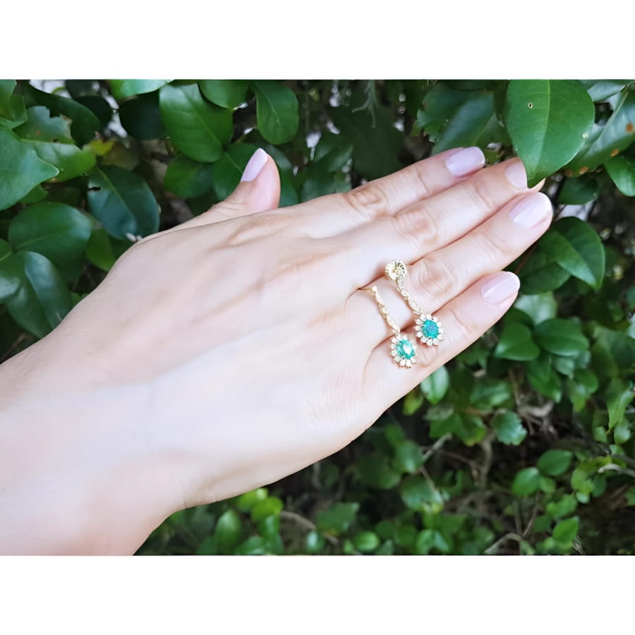 14K yellow gold dangle earrings with oval emeralds and halo diamonds on a hand.