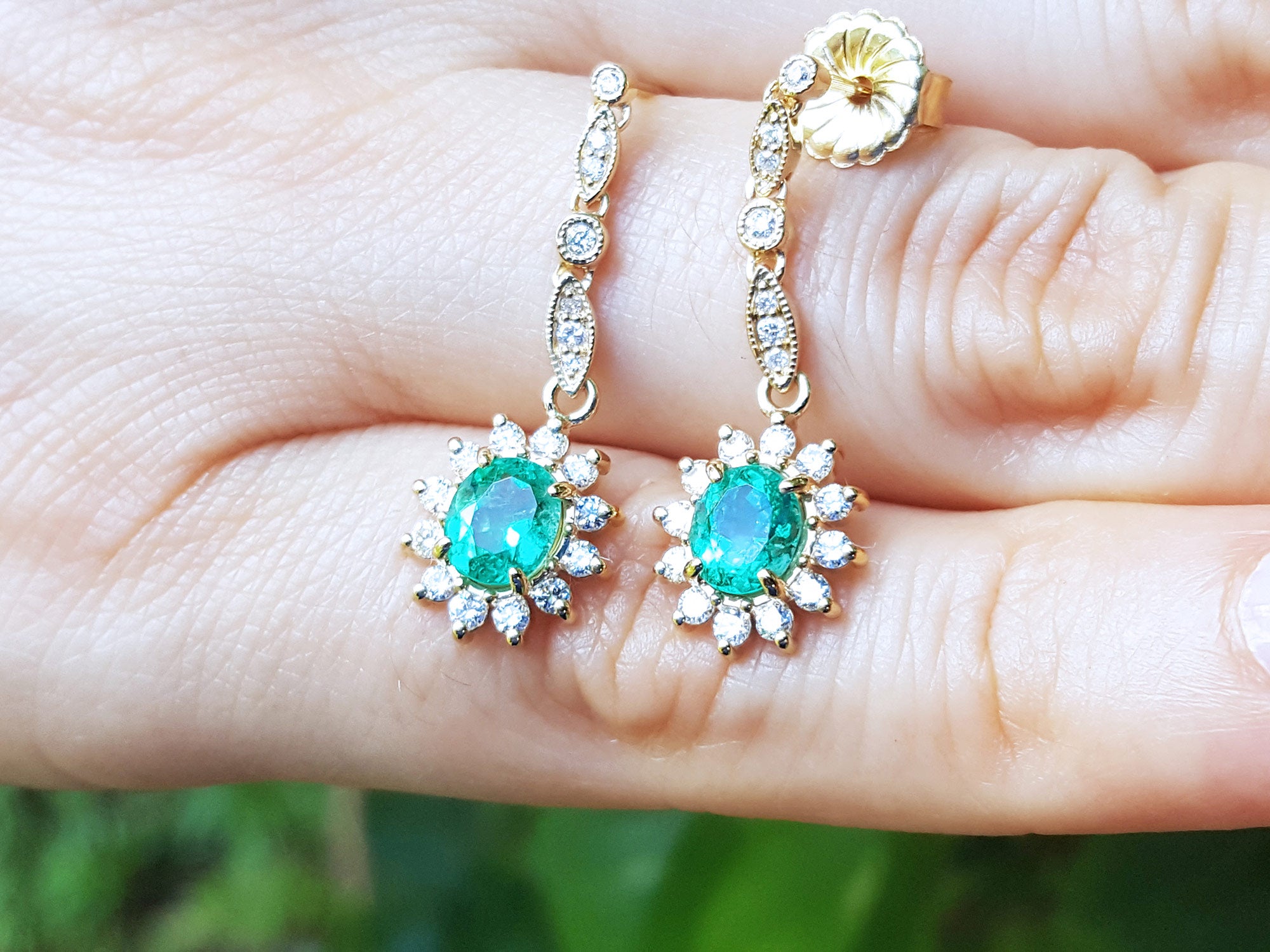 Gold earrings with emerald stones held in a hand against a blurred green background