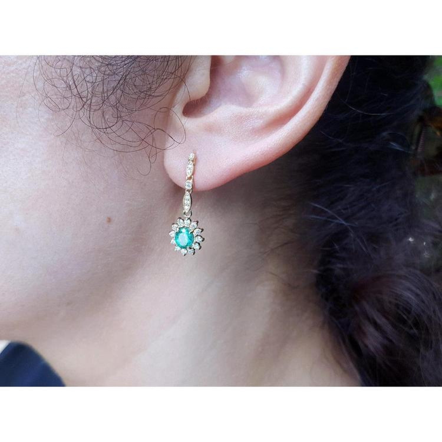 Oval emerald and halo diamond dangle earrings in 14K yellow gold on an ear.
