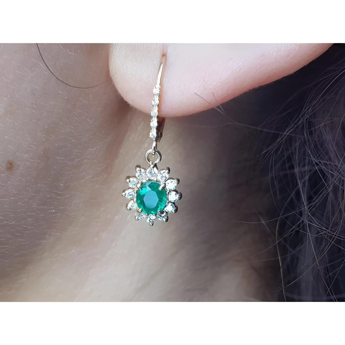 Close-up of a halo diamond oval cut emerald earrings lever backs worn on a woman's ear.