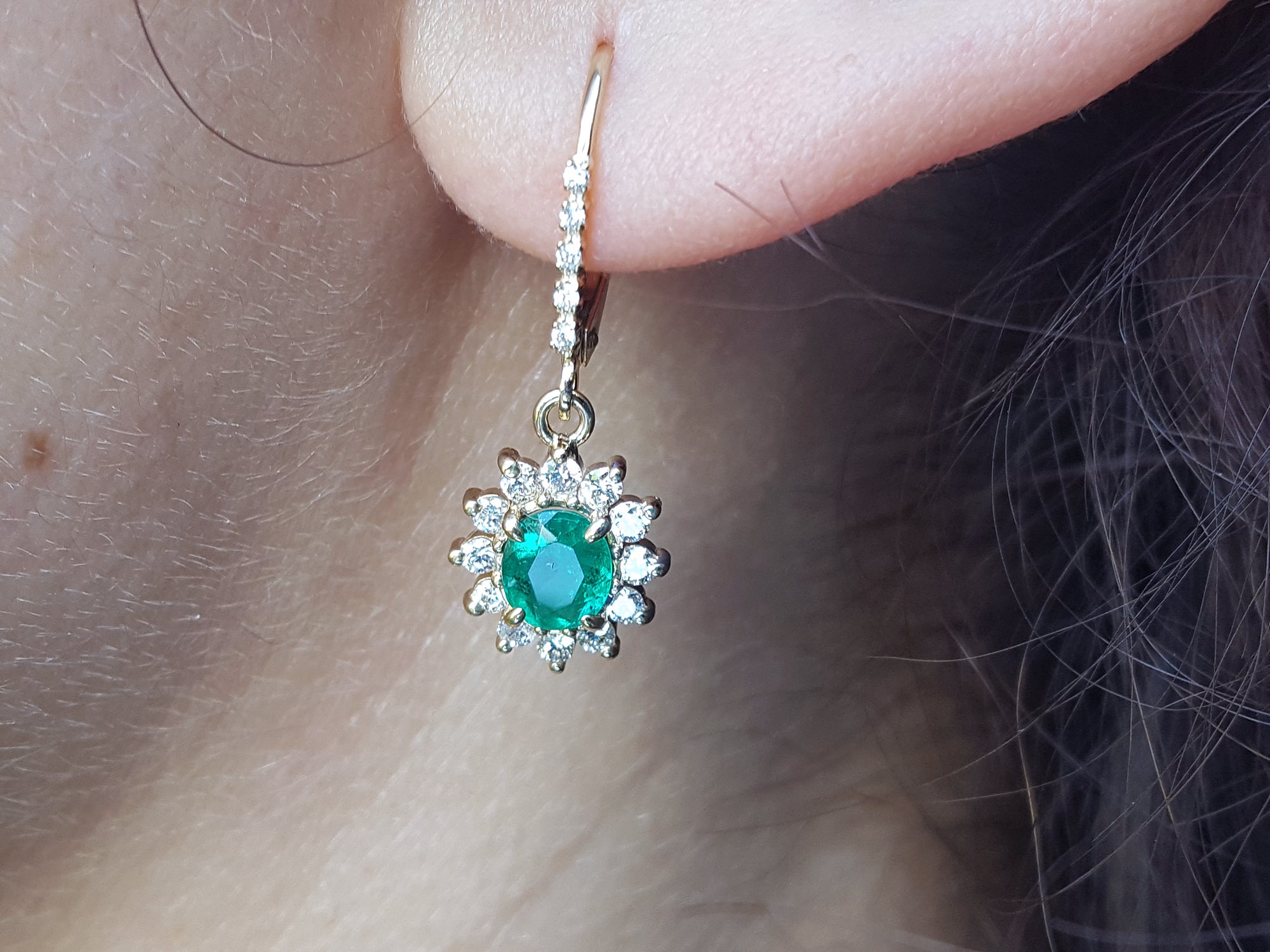 Close-up of a halo diamond oval cut emerald earrings lever backs worn on a woman's ear.