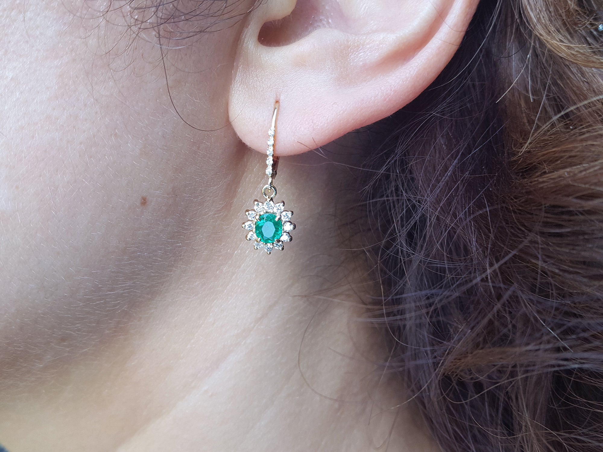 Close-up of a woman wearing a natural oval cut emerald earring, with halo diamond, yellow gold.