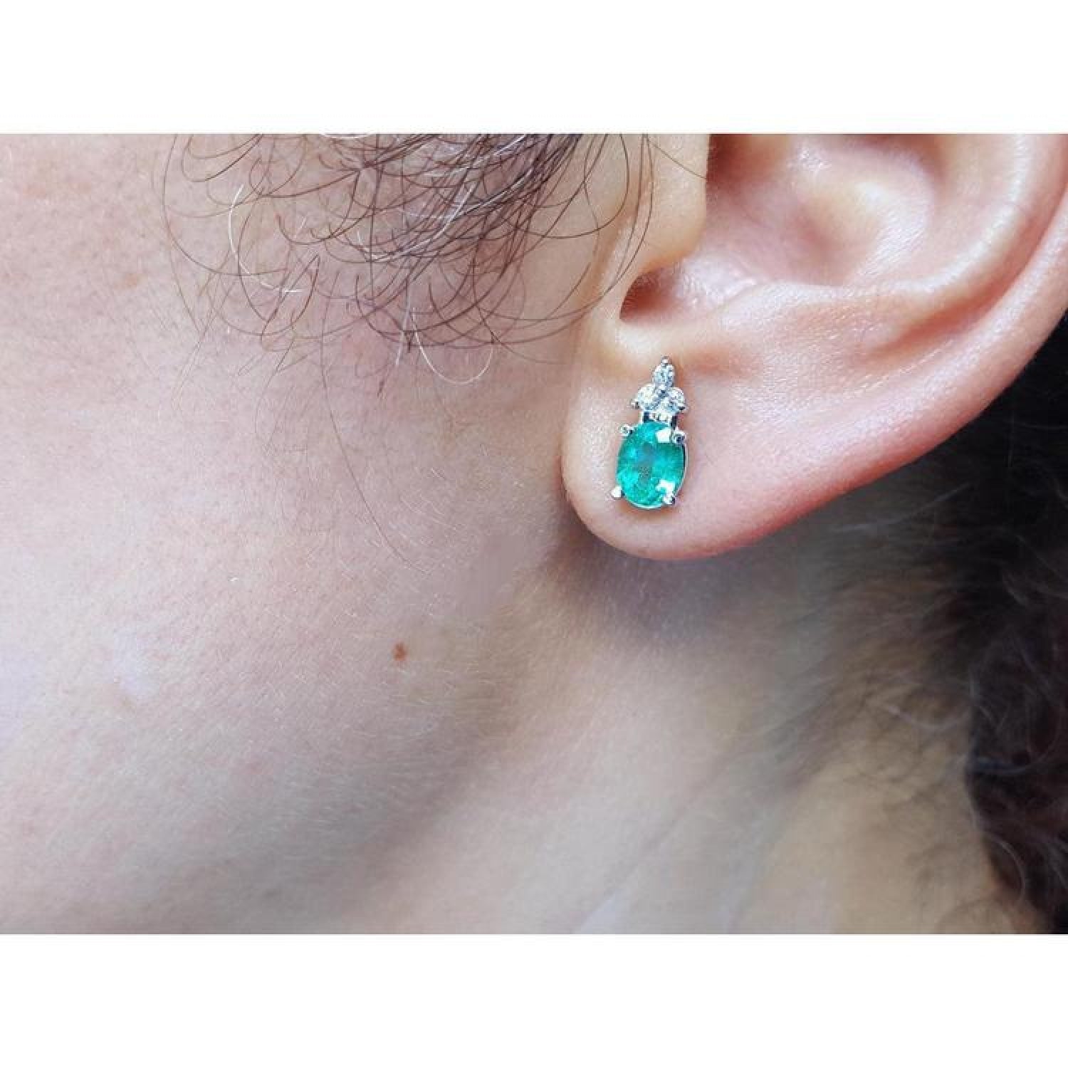 Oval cut natural emerald earrings with three diamonds in 14K white gold on an ear.