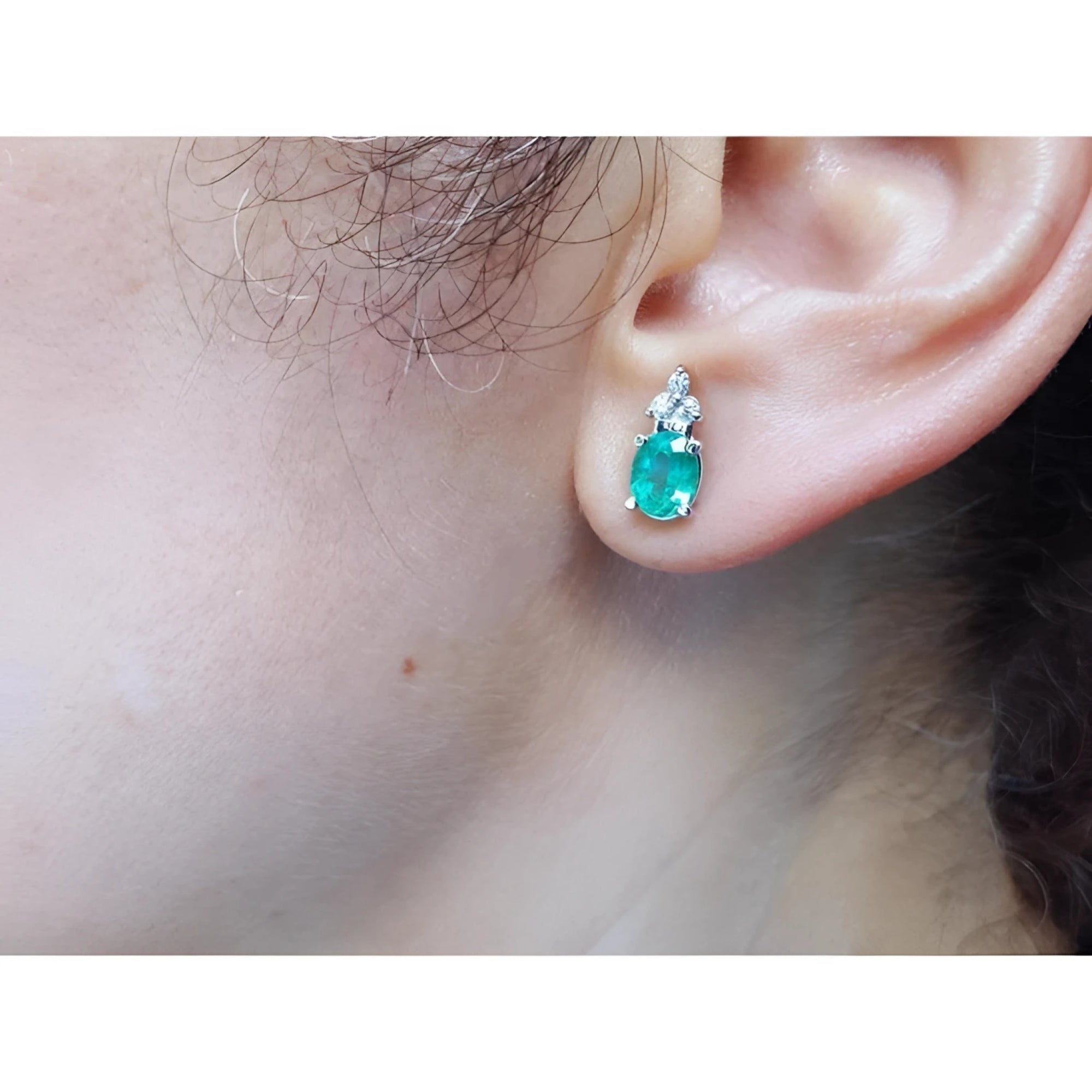 Oval cut natural emerald earrings with three diamonds in 14K white gold on an ear.