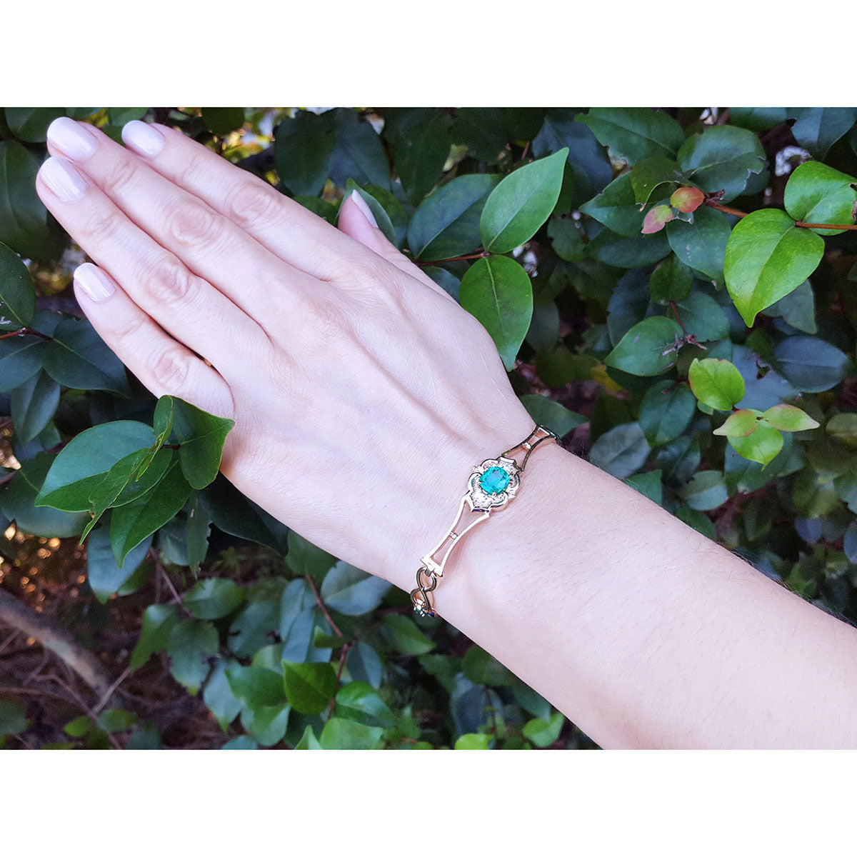 Hand wearing a gold bracelet with a natural emerald against a green leafy background.