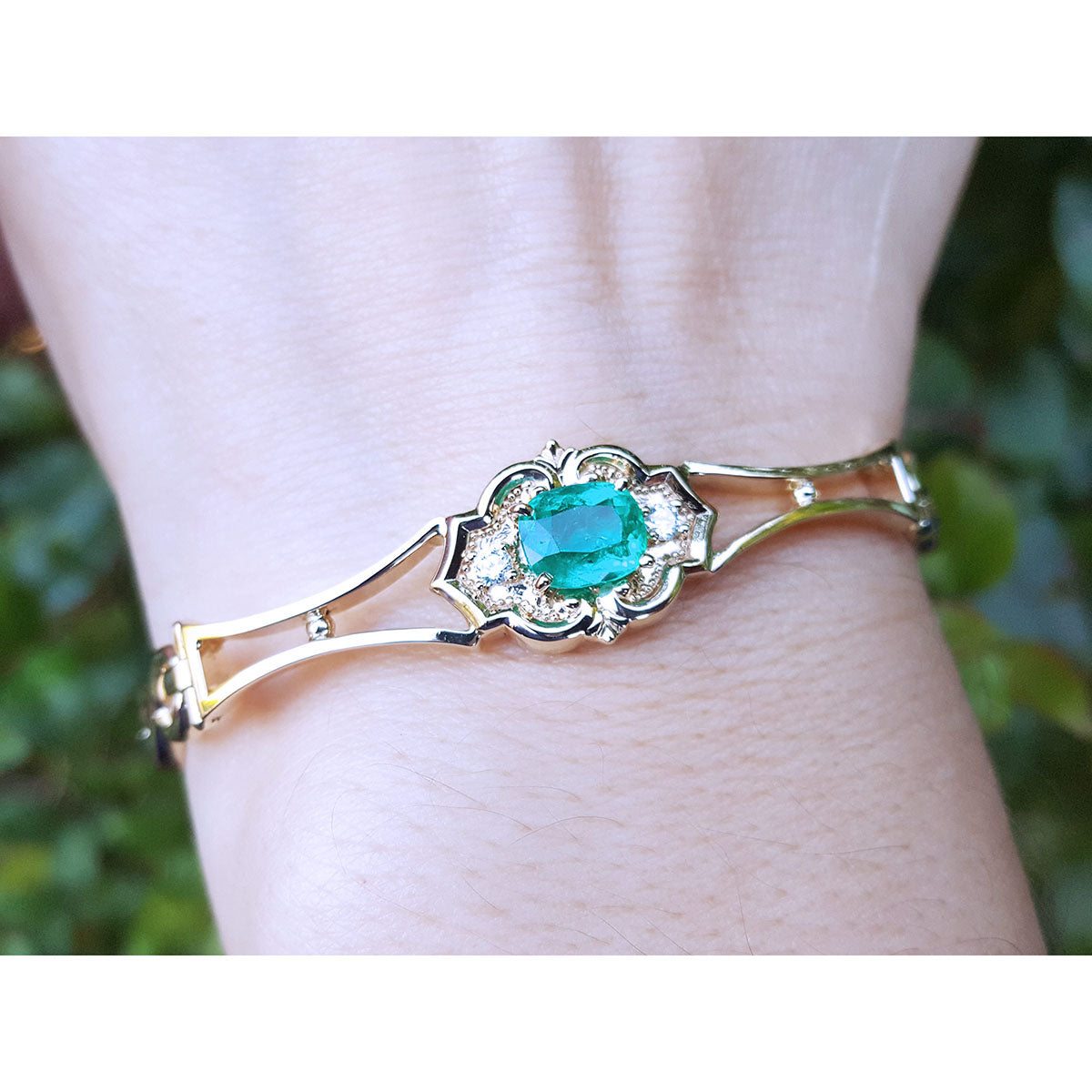 Gold bracelet with a natural colombian emerald oval cut on a wrist against a blurred green background.