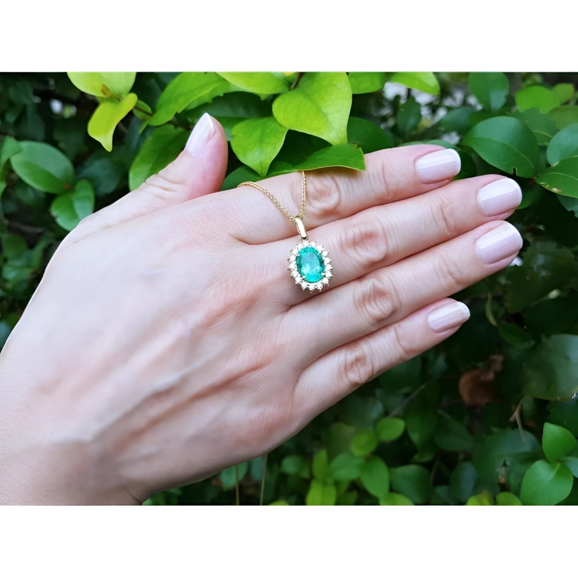 Oval natural emerald pendant with halo diamonds in 18K yellow gold, worn on a hand.