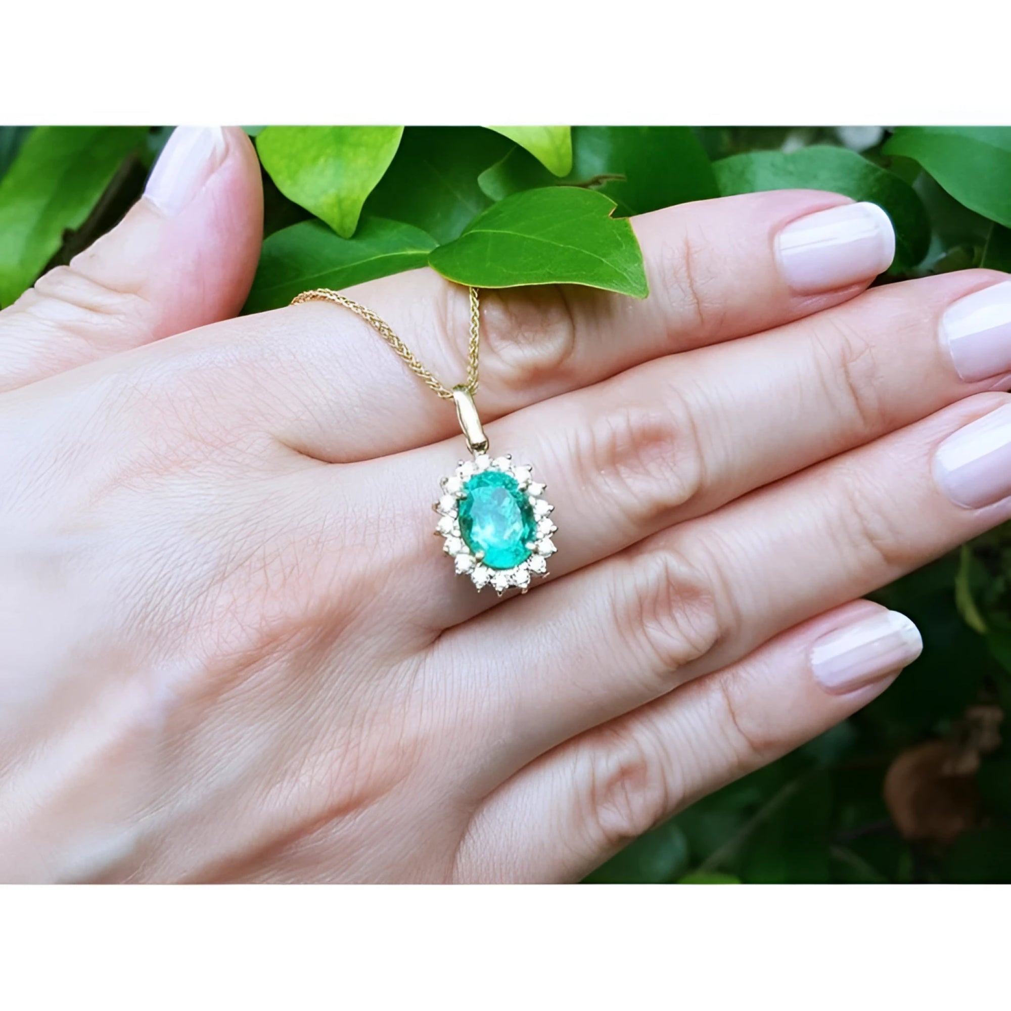 Oval natural emerald pendant with halo diamonds in 18K yellow gold, held in a hand.