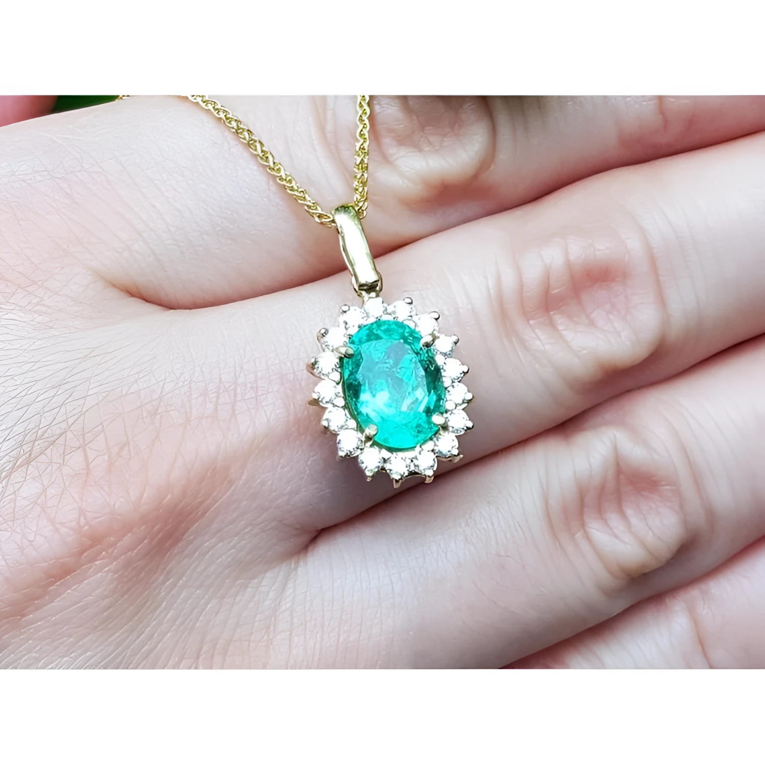 Oval natural emerald pendant with halo diamonds in 18K yellow gold, worn on a hand.