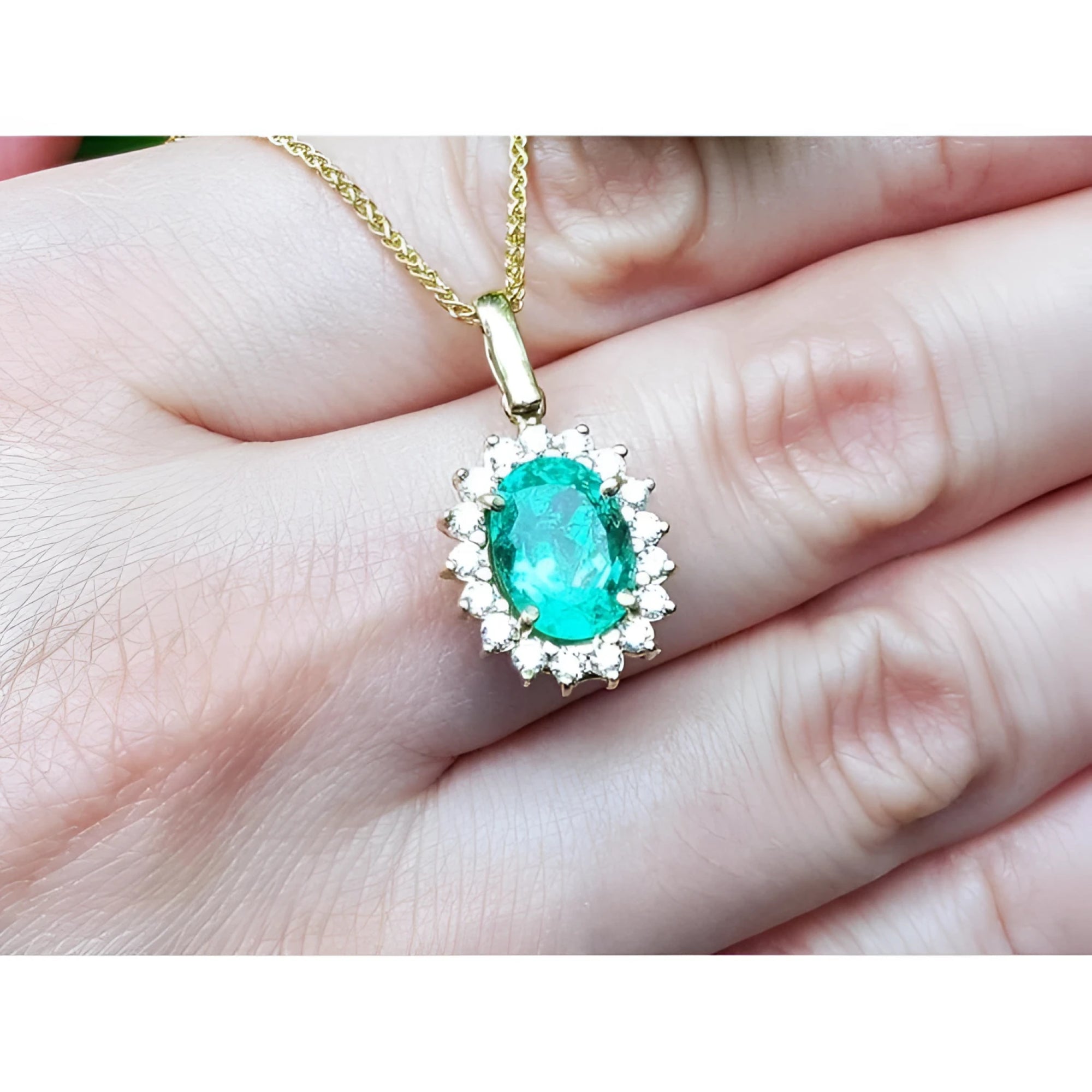 Oval natural emerald pendant with halo diamonds in 18K yellow gold, worn on a hand.