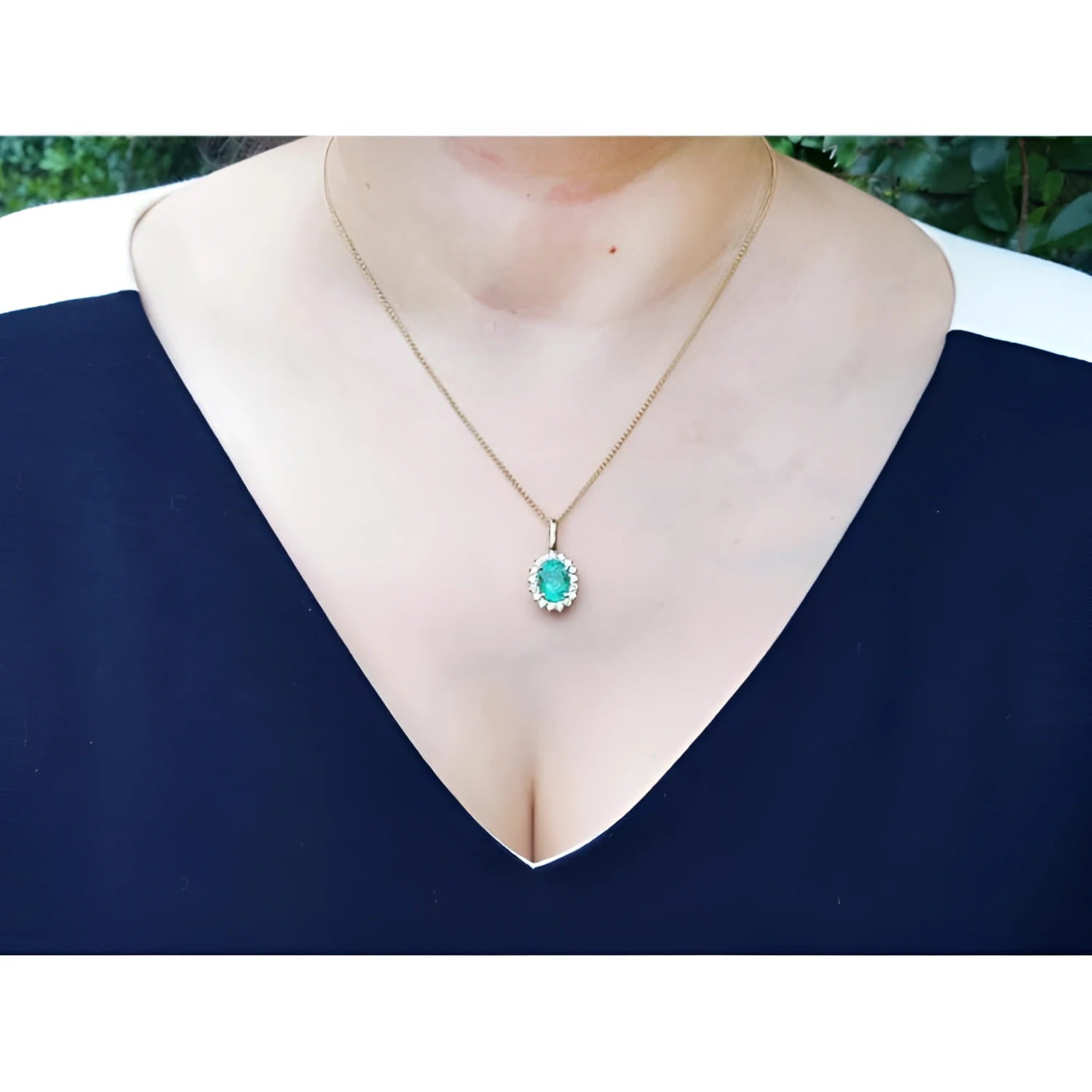 Oval natural emerald pendant with halo diamonds in 18K yellow gold, worn on a woman's neck.