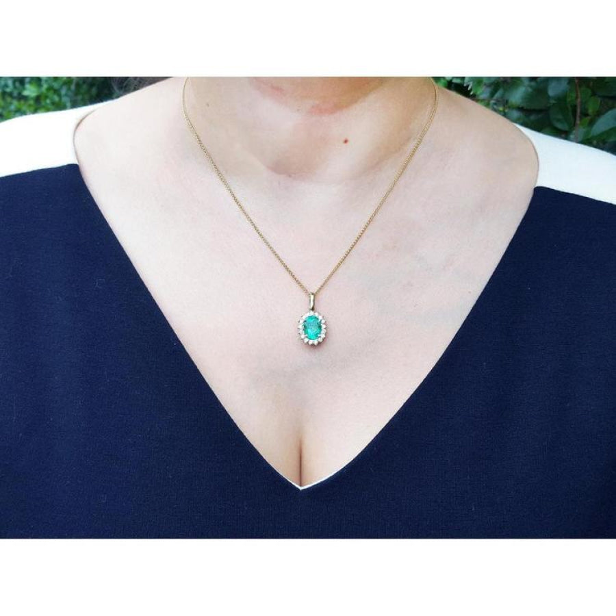 Oval natural emerald pendant with halo diamonds in 18K yellow gold, worn on a woman's neck.