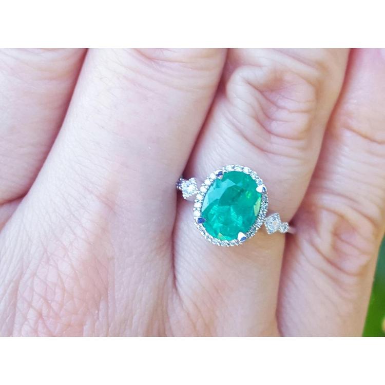 An oval cut emerald ring with accent diamonds on a hand with a blurred background.