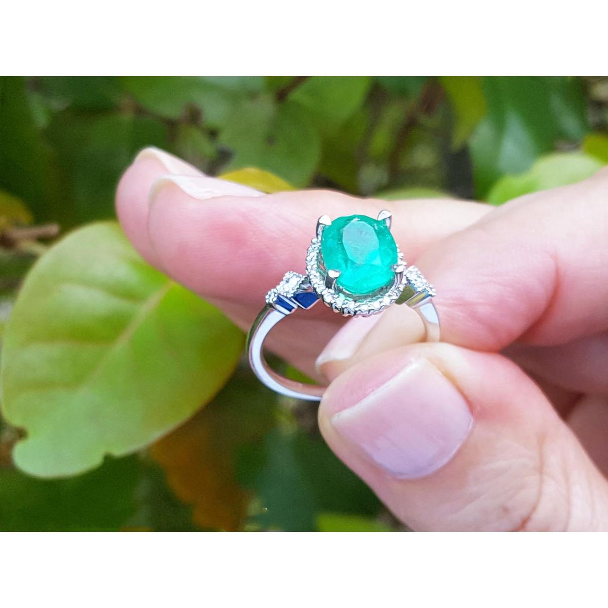 A white gold ring with n oval cut emerald held between fingers against a green leafy background.