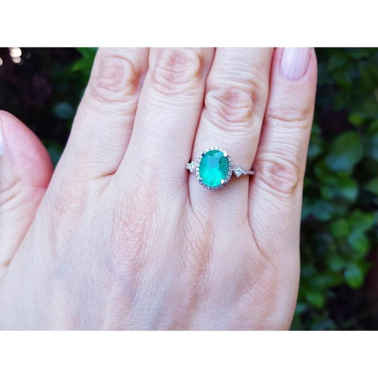 Oval-cut 2.02 ct. deep green emerald halo ring with diamonds on a 14K white gold band, worn on a hand.
