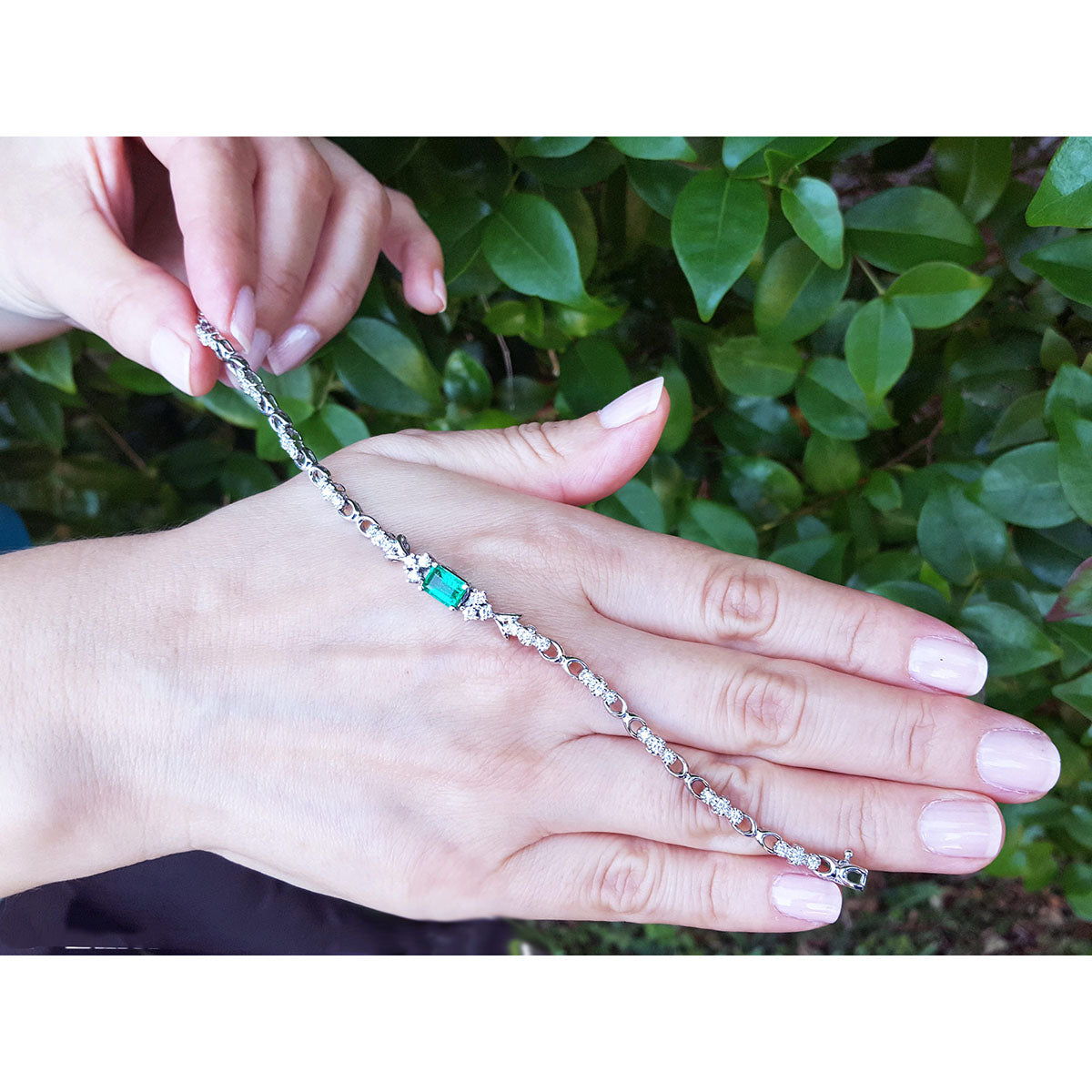 On hand displaying a bracelet with a center natural emerald against a green leafy background.