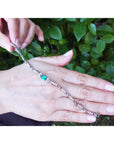 On hand displaying a bracelet with a center natural emerald against a green leafy background.