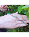 Hand showing a white gold bracelet with round emeralds against a green leafy background.