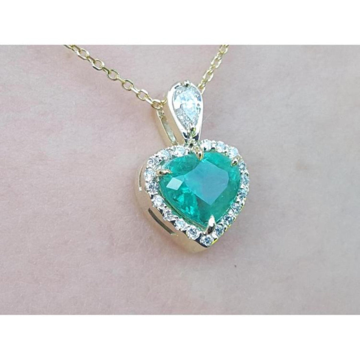 Heart-shaped emerald pendant with diamond accents on a gold chain against a light pink background