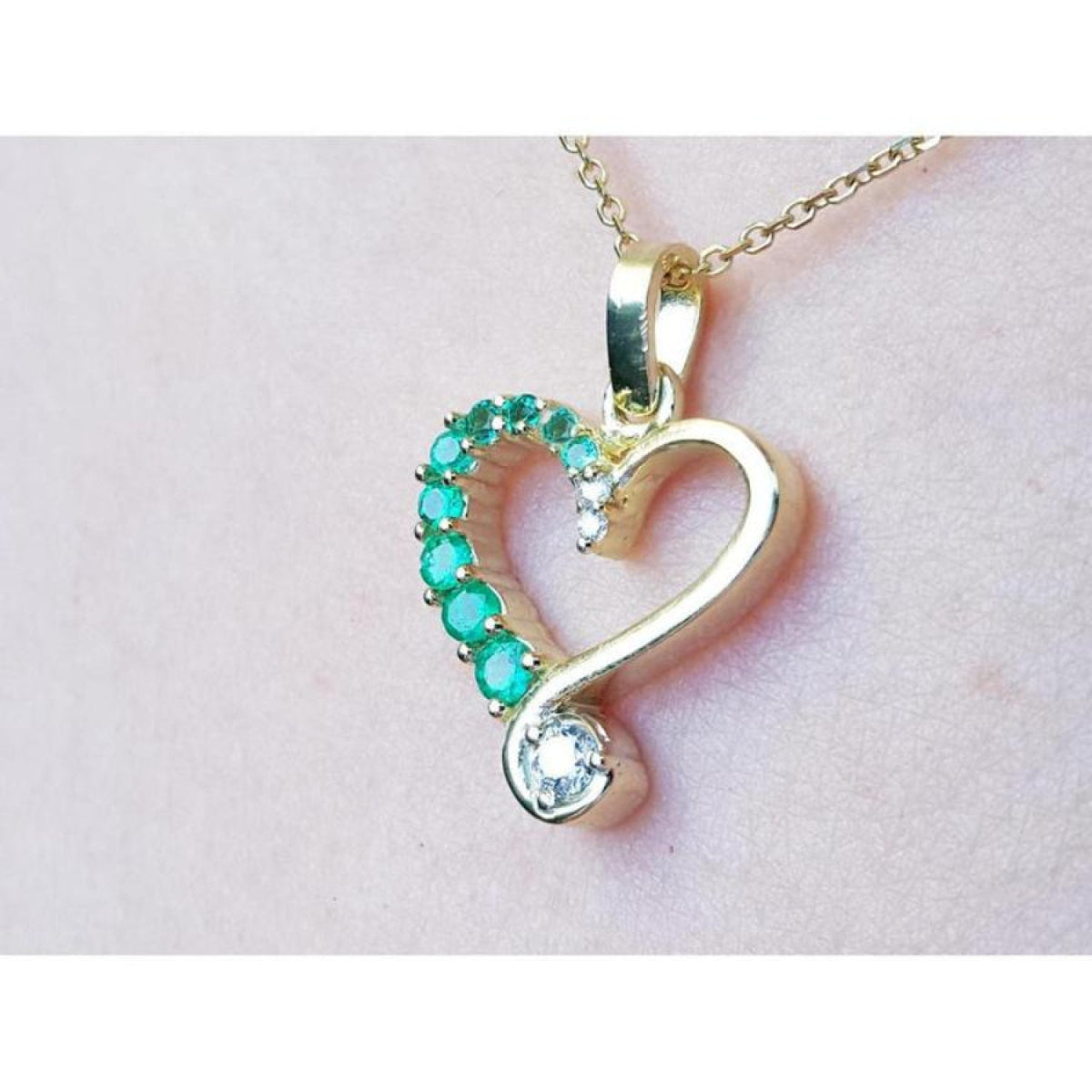 Heart-shaped gold pendant with emerald gemstones on a light pink background.