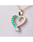 Heart-shaped gold pendant with emerald gemstones on a light pink background.
