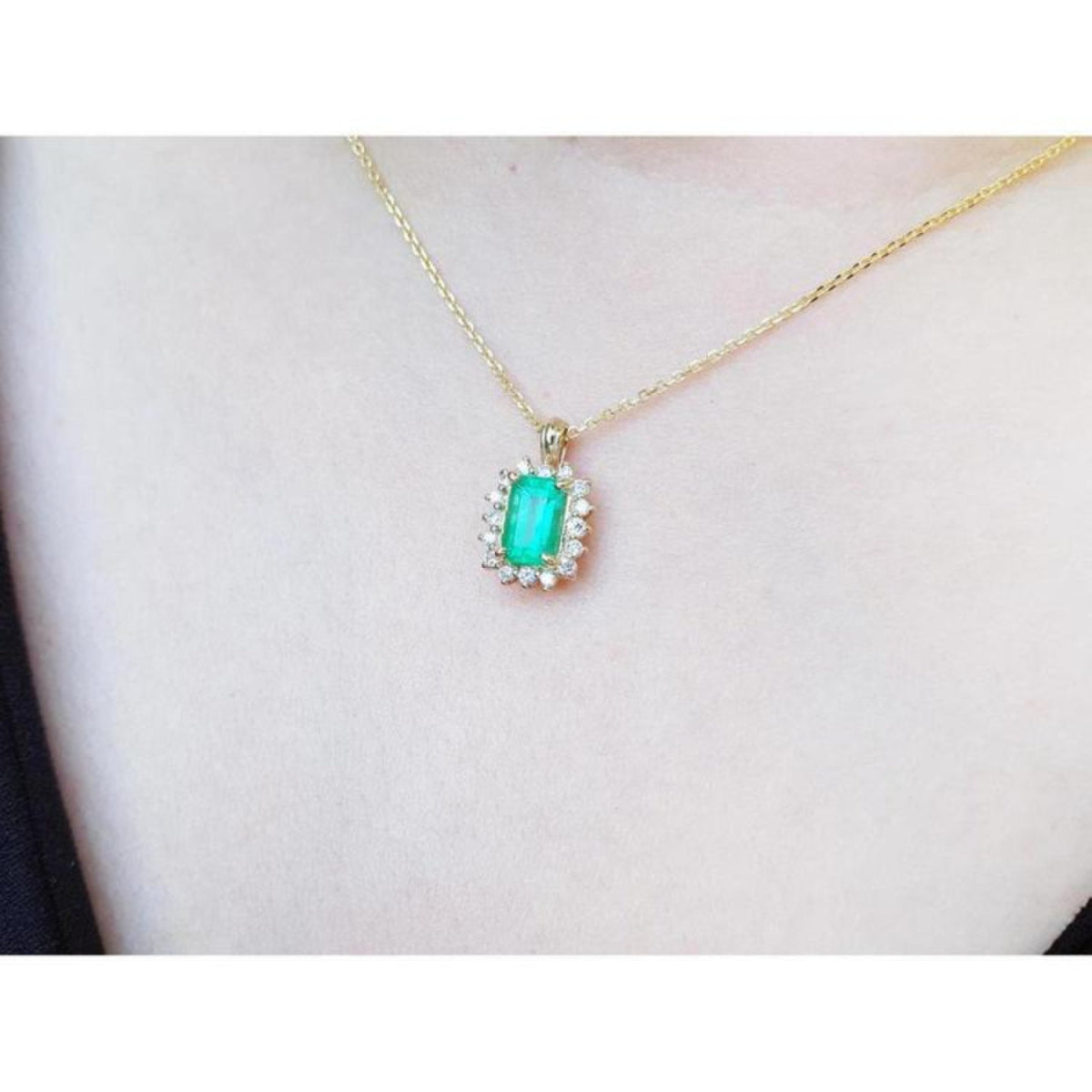 Emerald pendant with a halo of diamonds on a gold chain, worn on a person's chest.