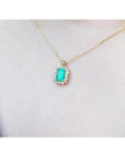 Emerald pendant with a halo of diamonds on a gold chain, worn on a person's chest.