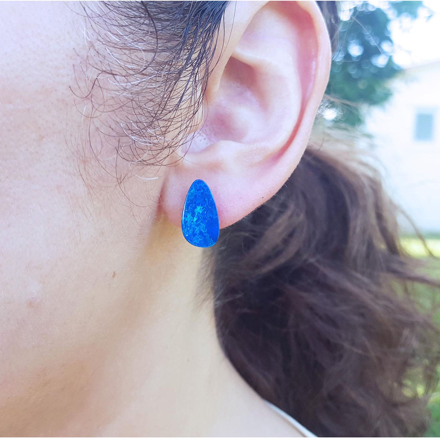 Close-up of an ear wearing a blue opal doublet for earring with a blurred outdoor background.