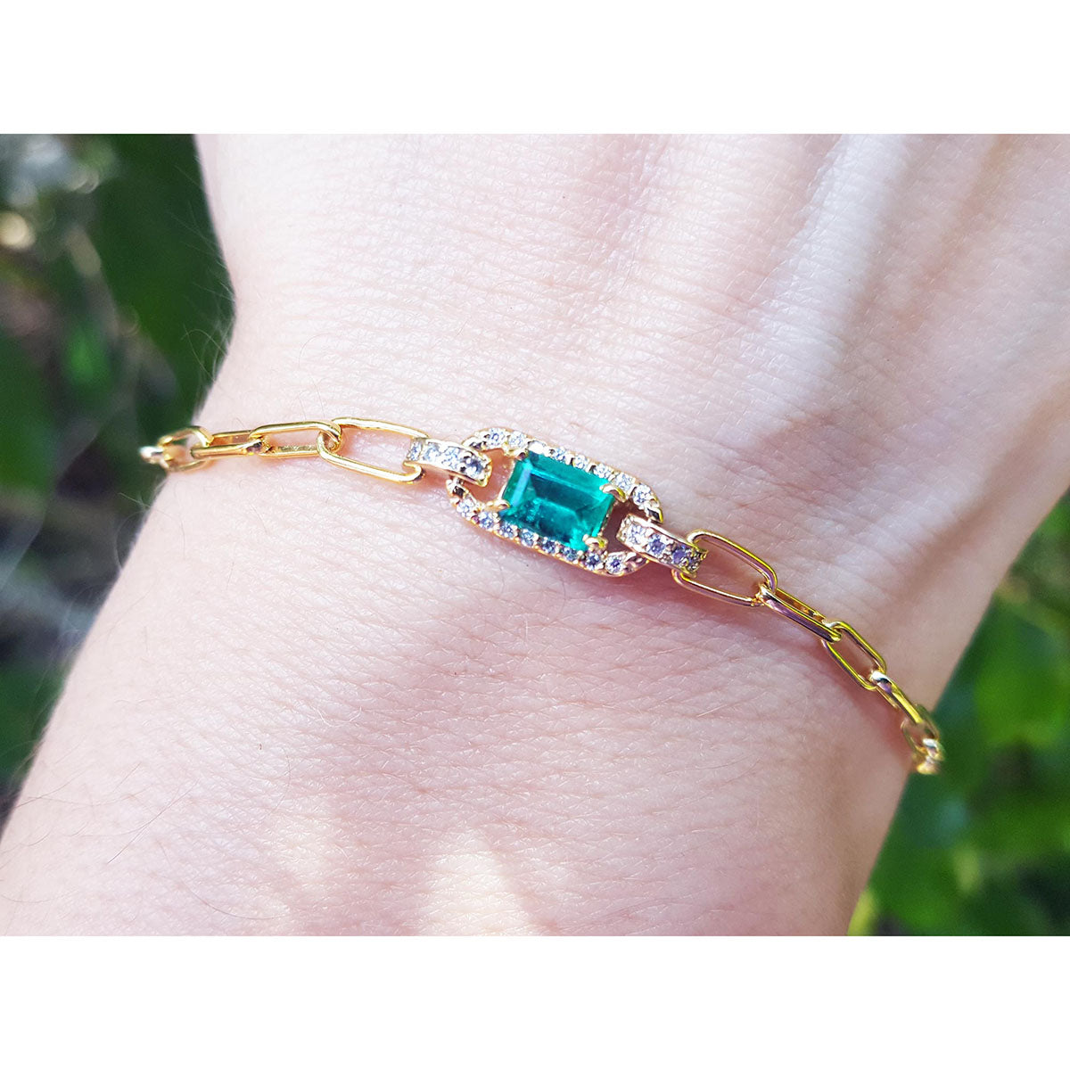 Gold paperclip bracelet with a natural emerald on a wrist against a blurred natural background.
