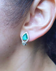 Close-up of an ear wearing an emerald teardrop earring with a blurred natural background